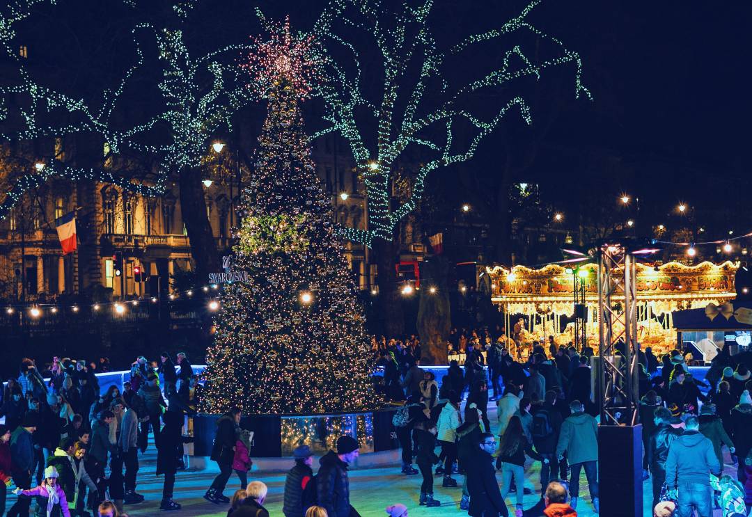 People ice skating at night on an open-air ice-rink surrounded by trees decorated for Christmas